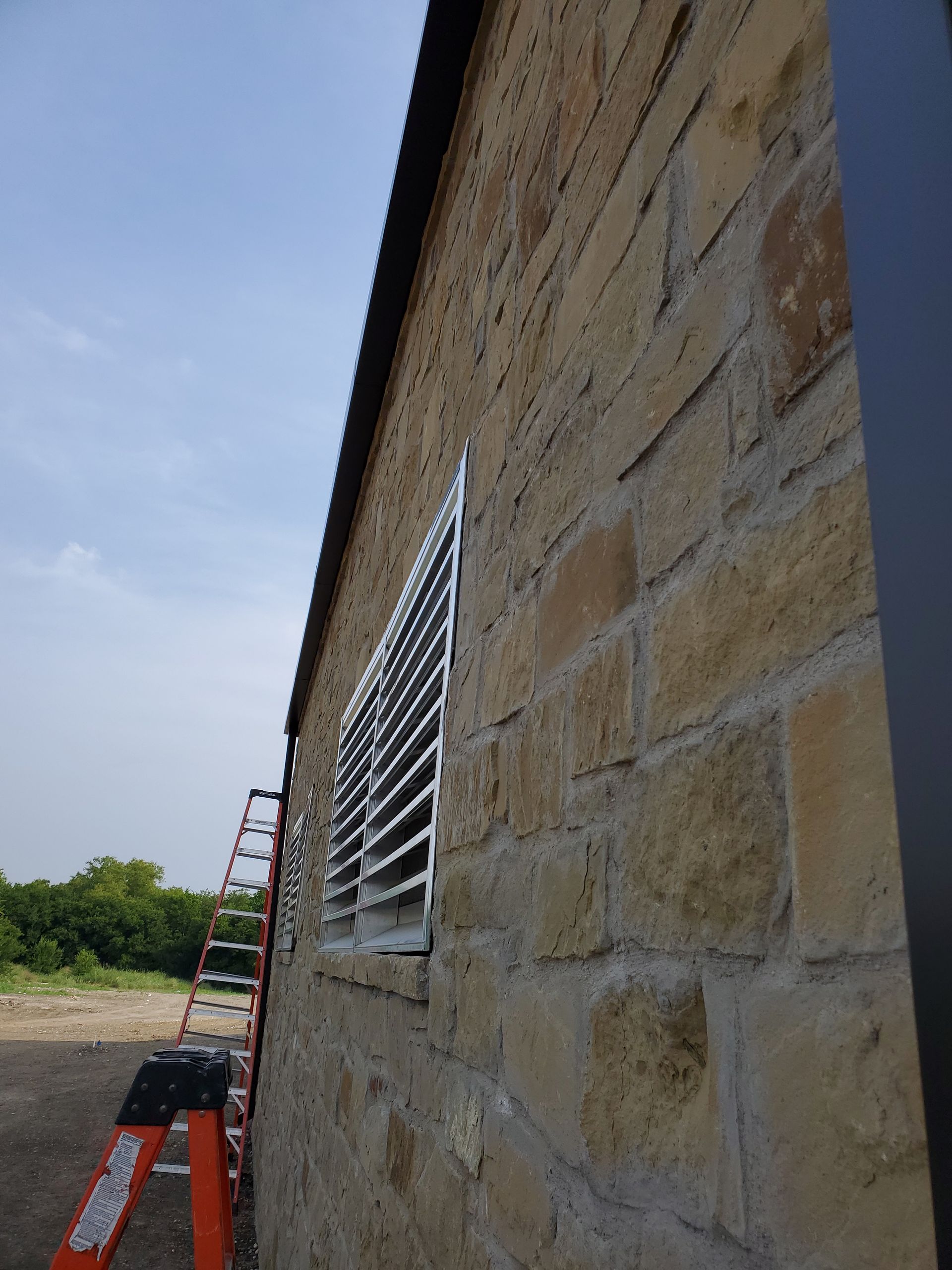 Stone building exterior with metal window grate; an orange ladder leans against the wall under a cloudy sky.