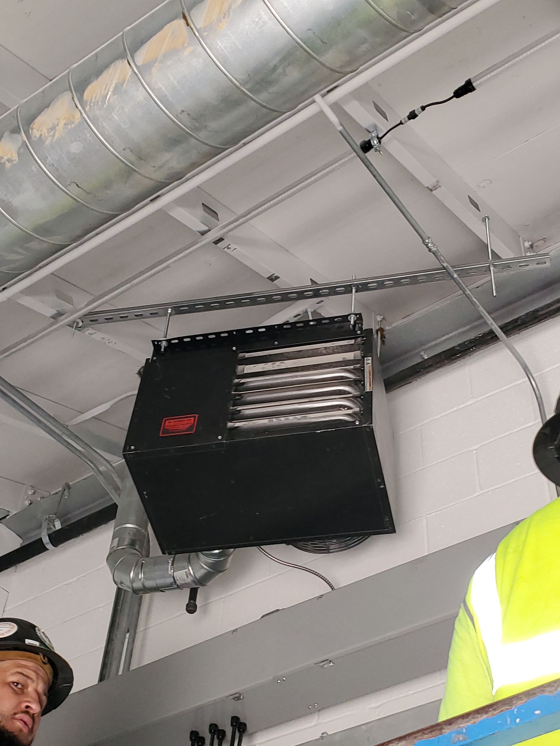 Black ceiling-mounted heater between HVAC ducting and fluorescent lighting, with two people in yellow vests nearby.