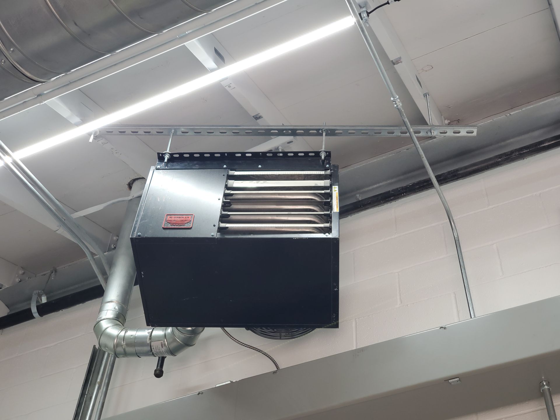 Black ceiling-mounted gas heater with exhaust ductwork. Mounted on a metal frame, near a white wall and ceiling.