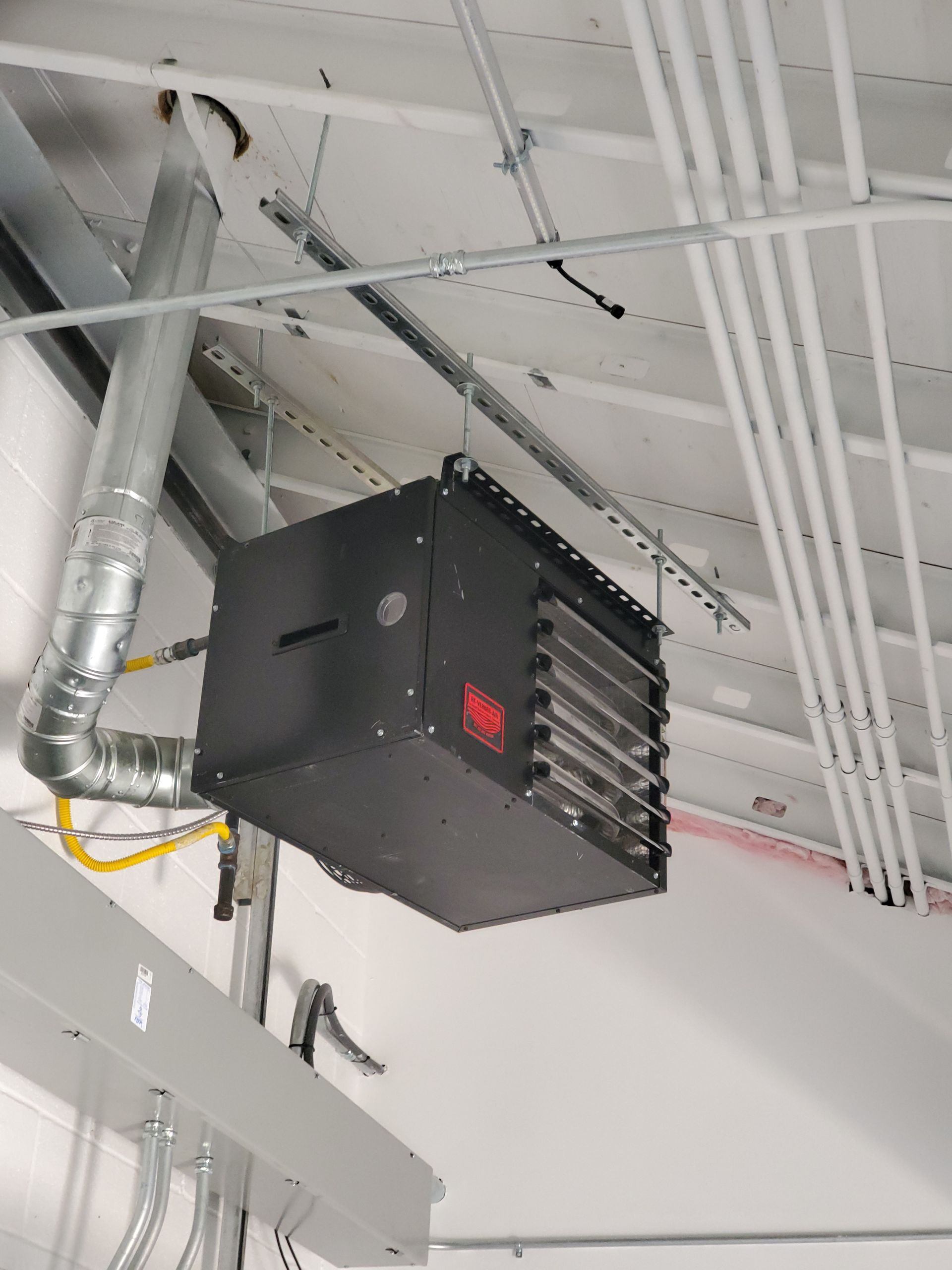 Black, rectangular industrial heater suspended from a ceiling, with ventilation and gas lines visible.