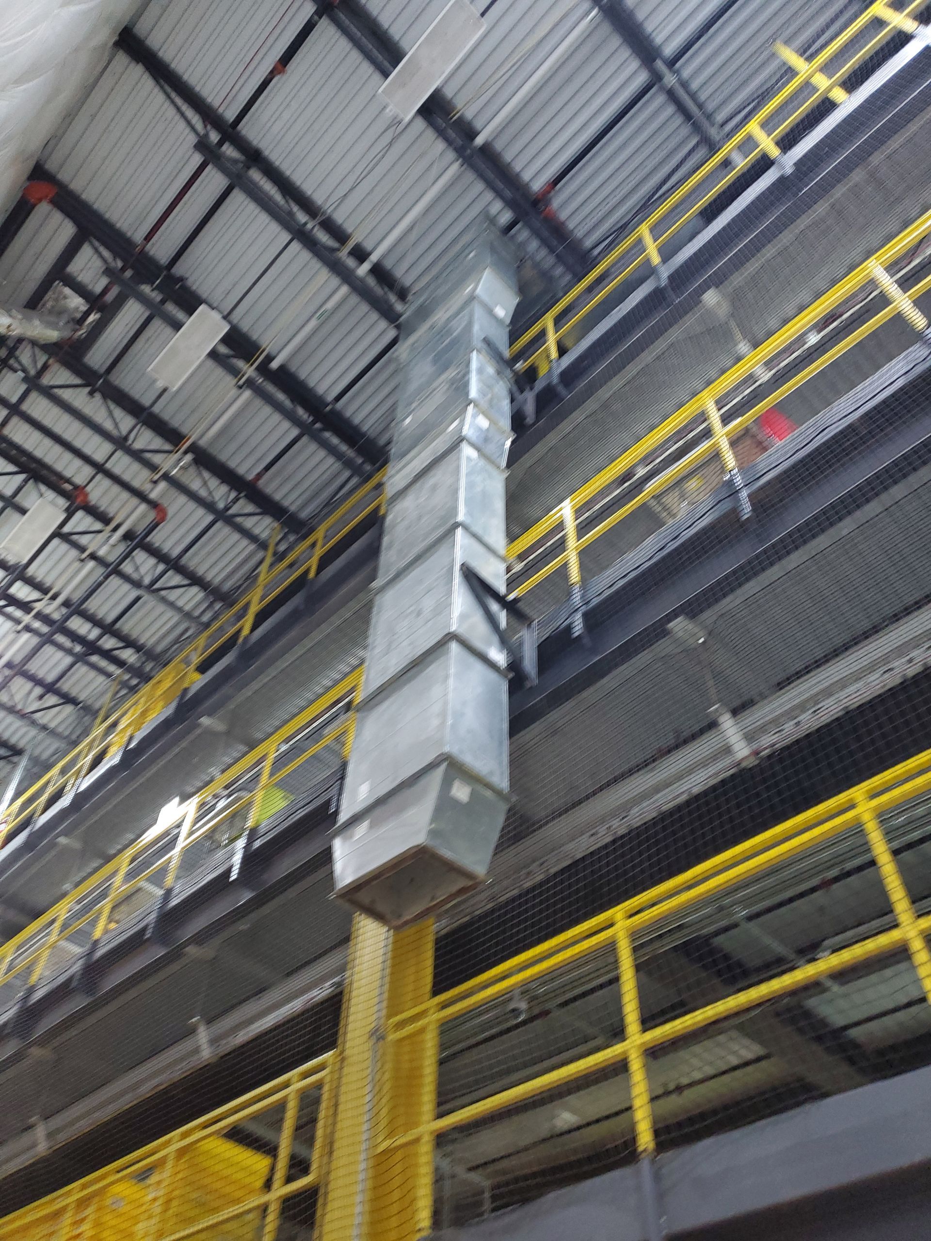 Metal ductwork suspended from a warehouse ceiling, running vertically near yellow safety railings.