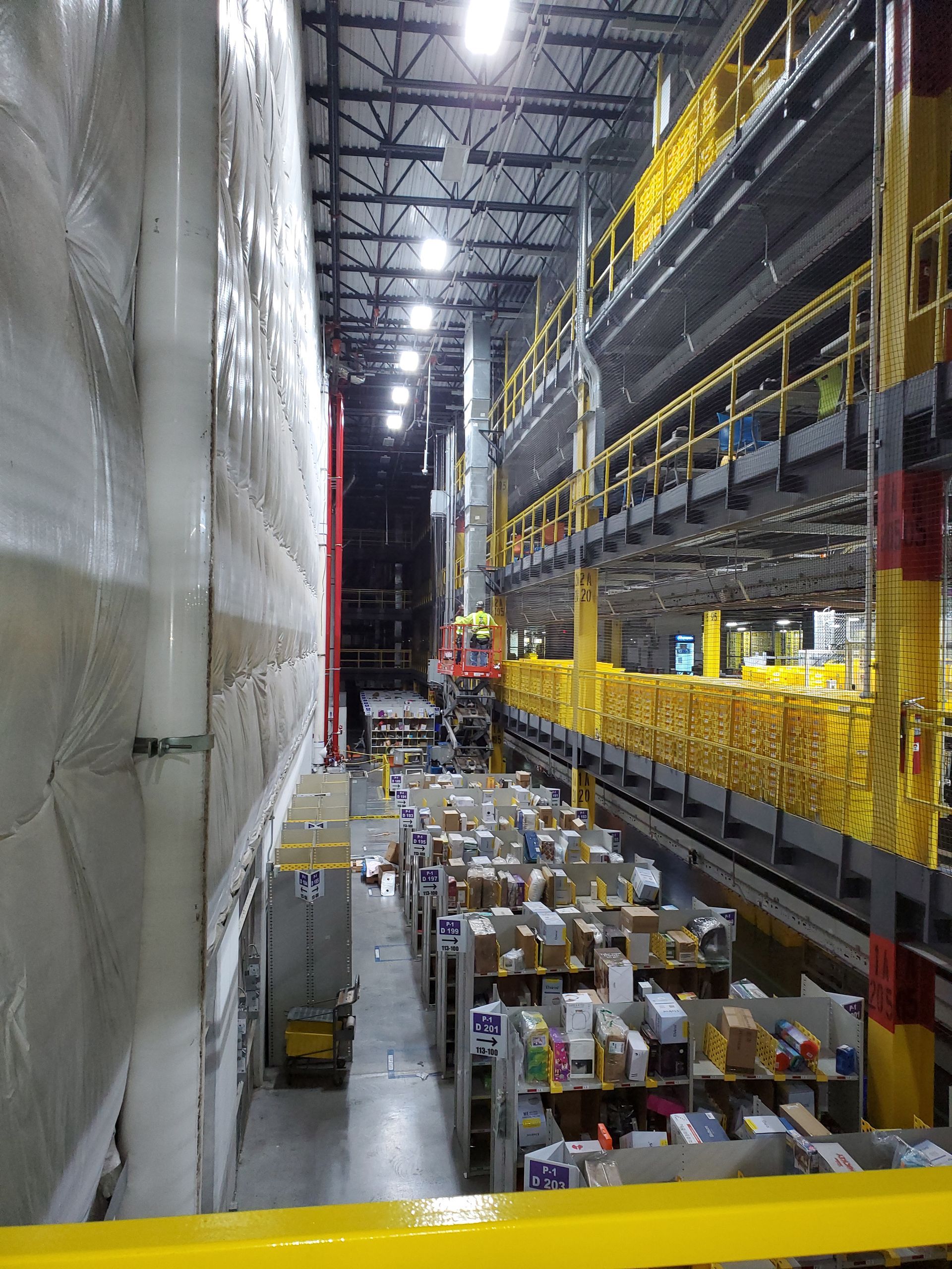 Warehouse interior with tall shelves, filled boxes, and elevated walkways. Overhead lighting.
