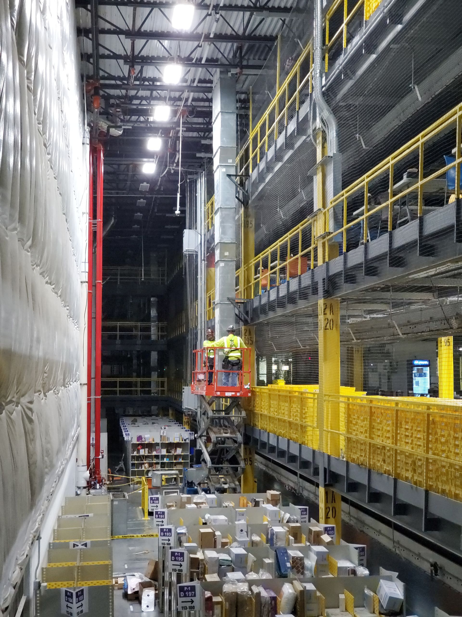 Warehouse interior with workers on lift; boxes on floor. Yellow safety railings, red lift, overhead lighting.