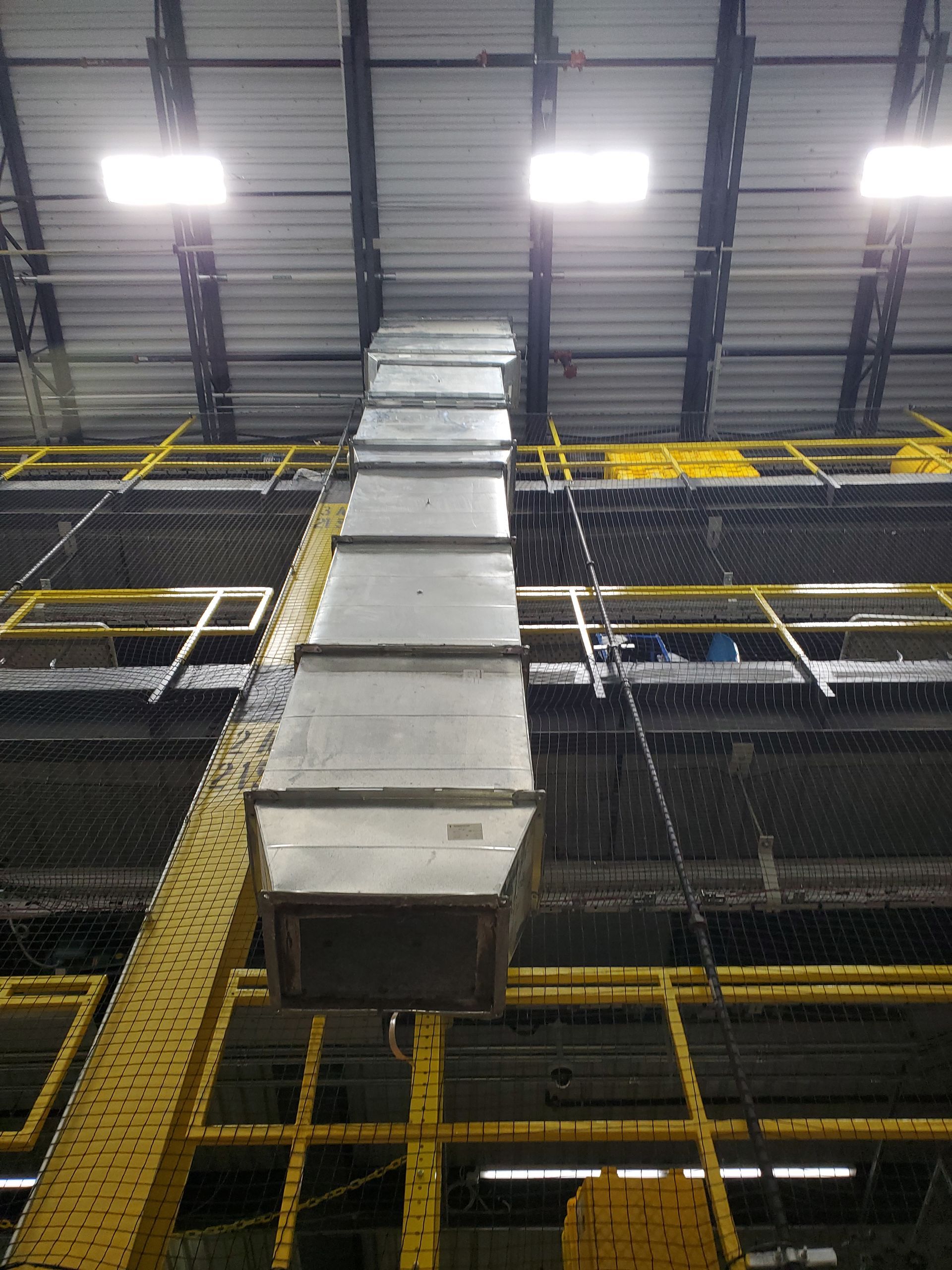 A metal ventilation duct suspended from a factory ceiling by wires. Yellow support structure visible.