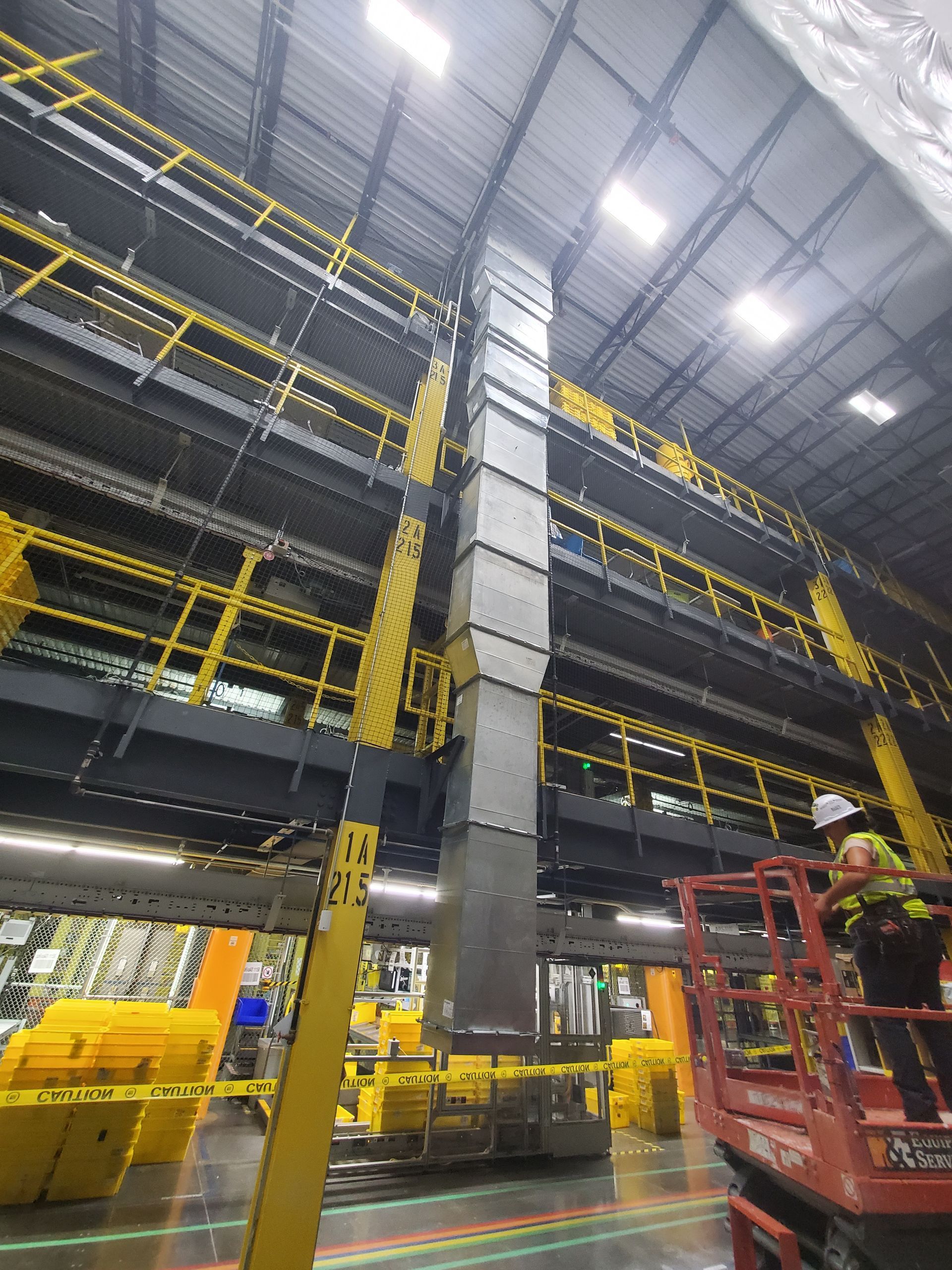 Warehouse interior with elevated walkways, a large vent, and worker on a lift. Yellow railings contrast with gray structure.