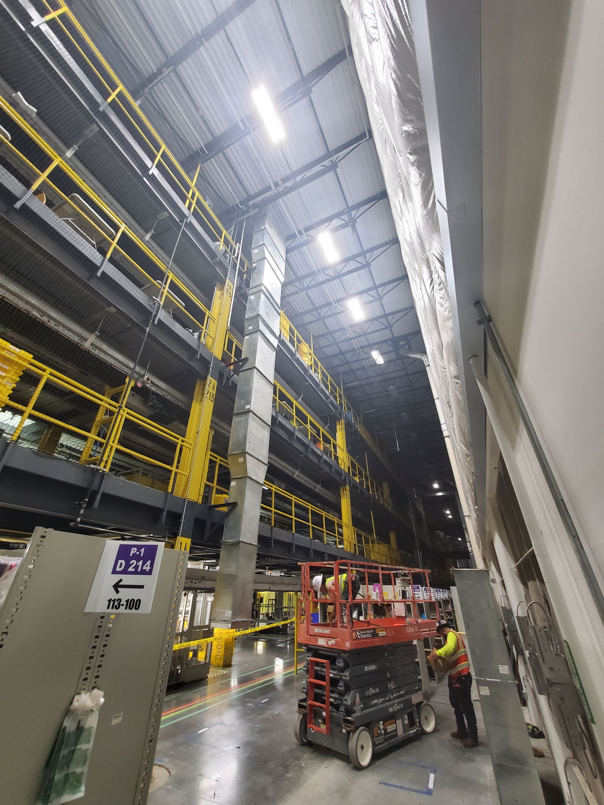 Inside a warehouse with tiered shelving, workers on a lift. Black, yellow, and gray interior.