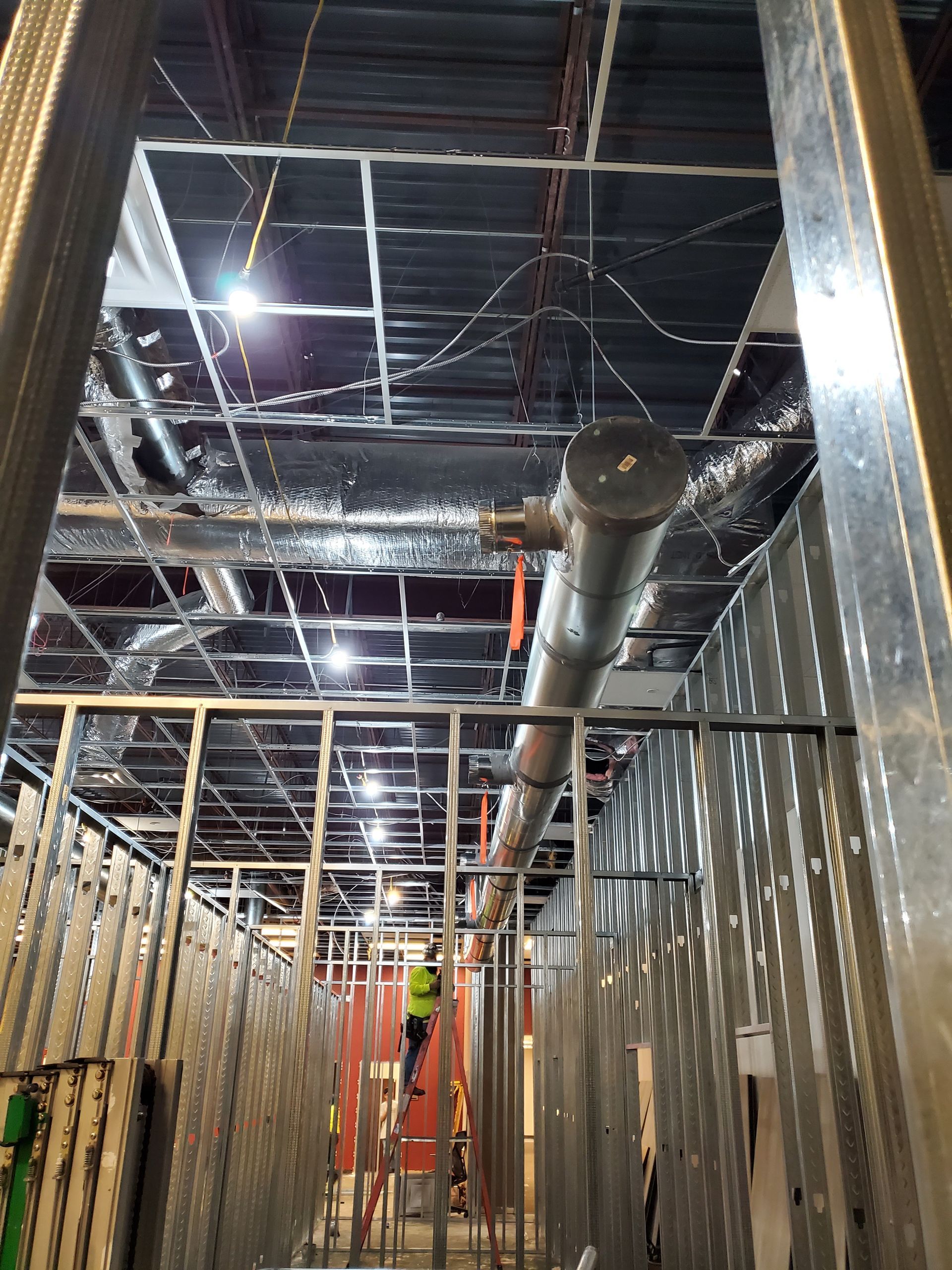 Construction site: metal framework, ductwork, and ceiling grid with a worker on a ladder.