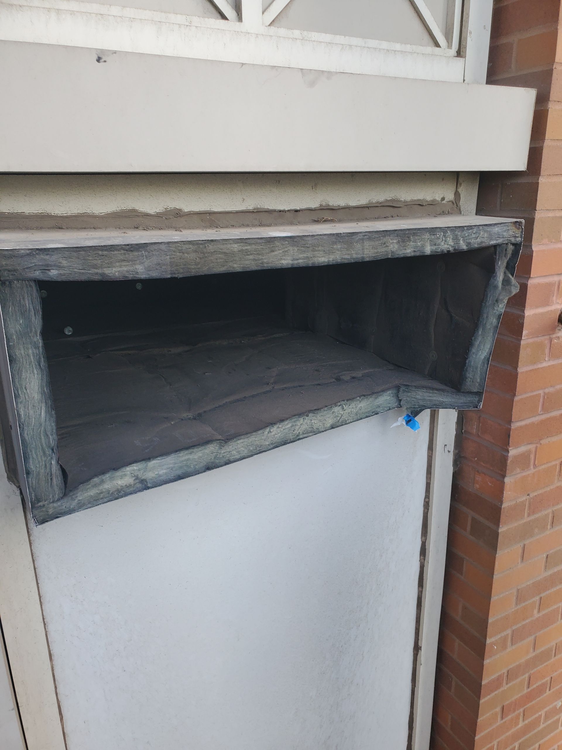 A rectangular air vent, grey with a dark interior, is installed above a white door, next to a brick wall.