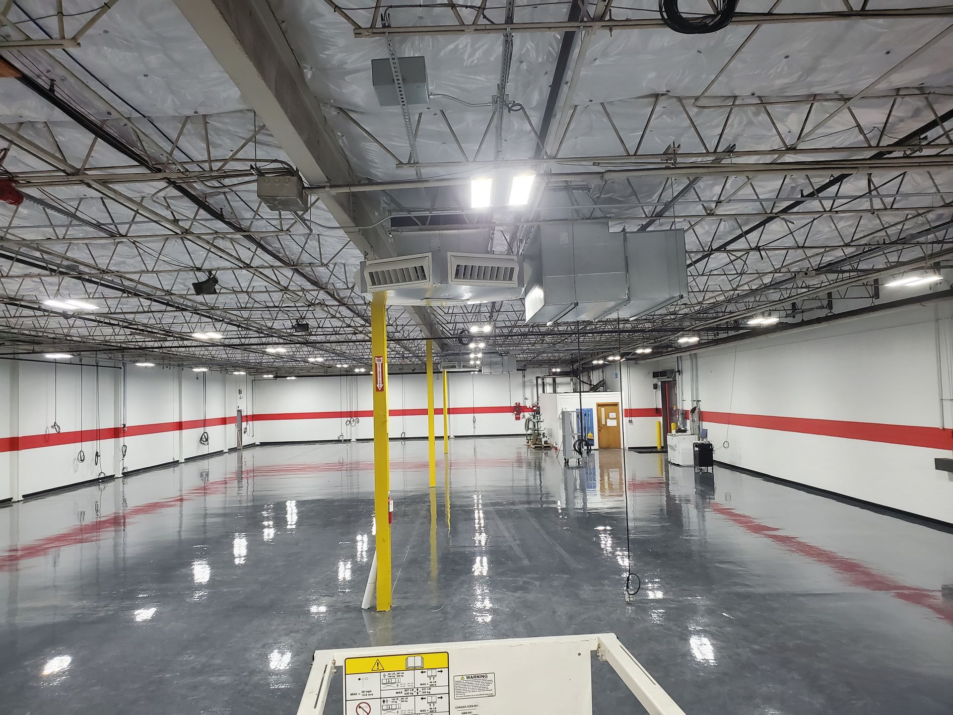Interior of a large, empty warehouse with a reflective floor, exposed ceiling, and red stripe along walls.