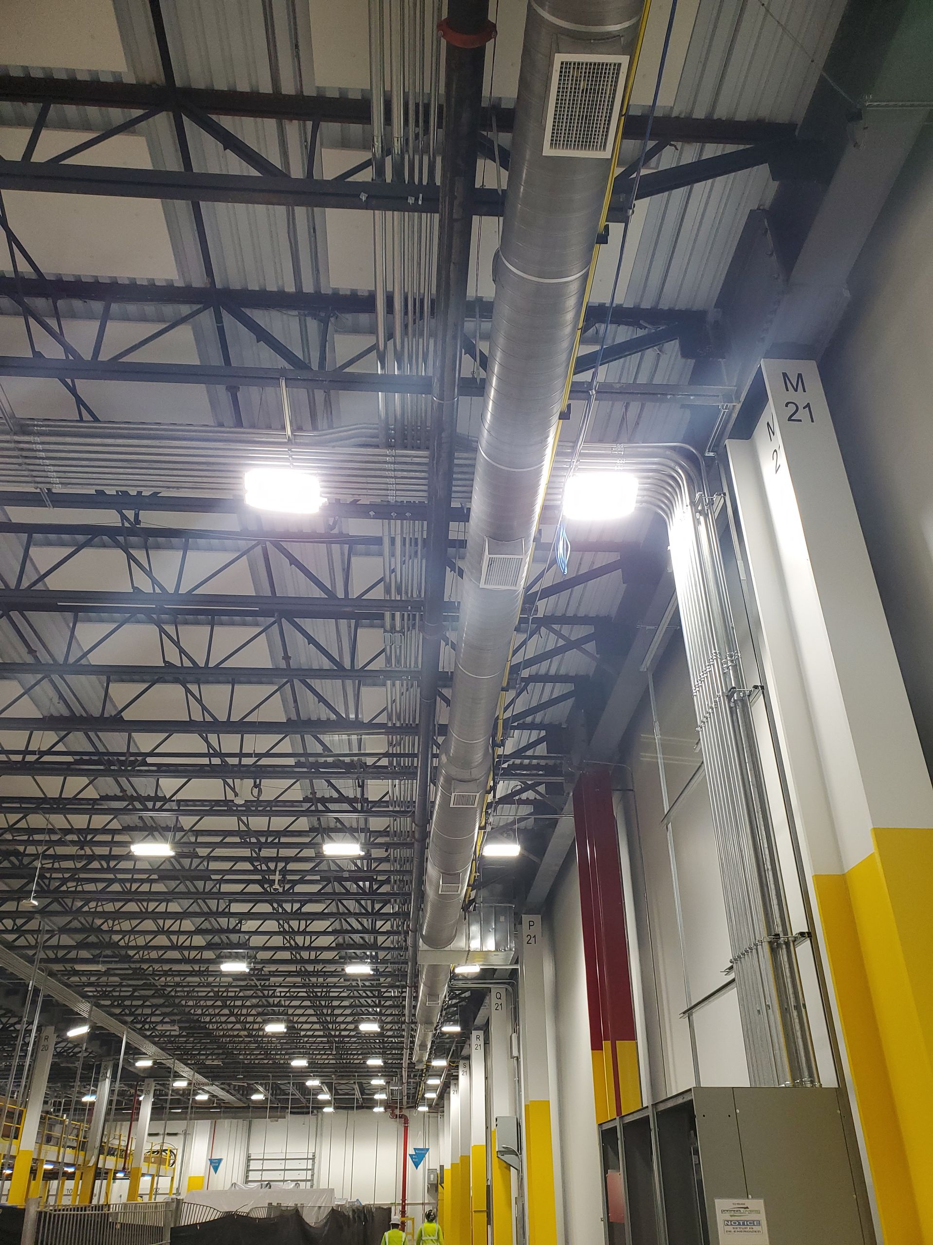 Warehouse ceiling with metal beams, ductwork, and lighting. Gray and yellow wall on right.