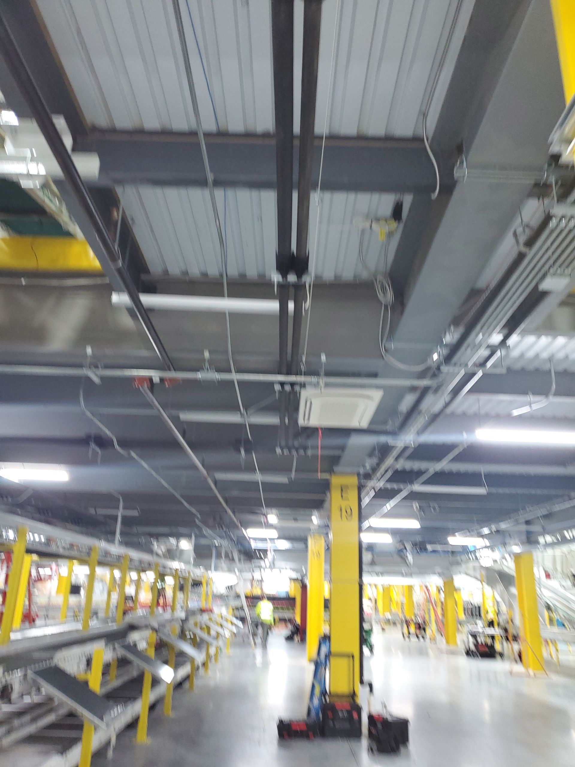 Interior of a warehouse with yellow support beams, gray ceiling with pipes and wiring, and conveyor belts.