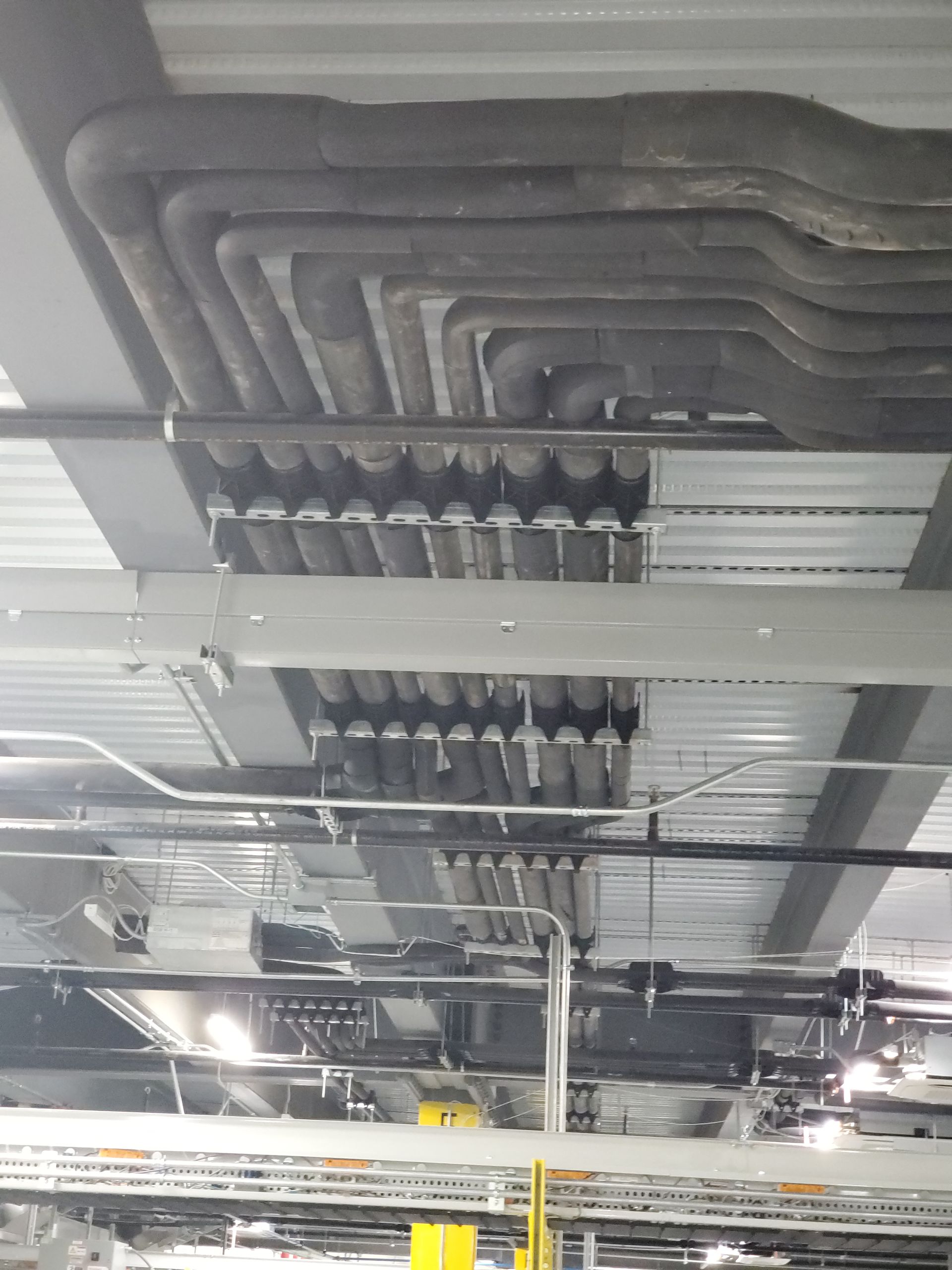 Overhead view of insulated pipes and conduits running across a metal ceiling in an industrial setting.