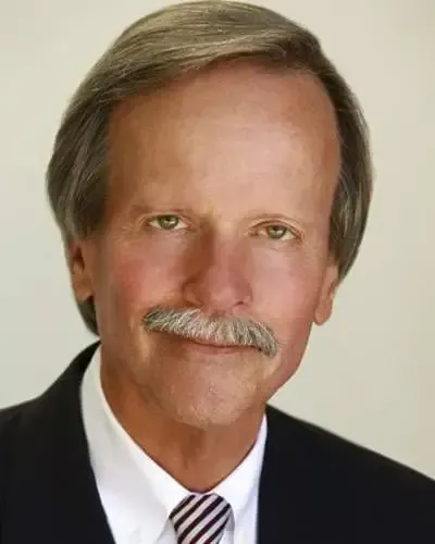 A man with graying hair and mustache in a suit smiles at the camera.
