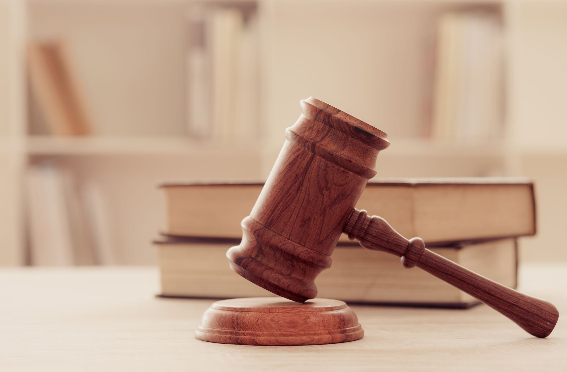 Wooden gavel on a stand in front of stacked books, on a table, with a blurred bookshelf backdrop.