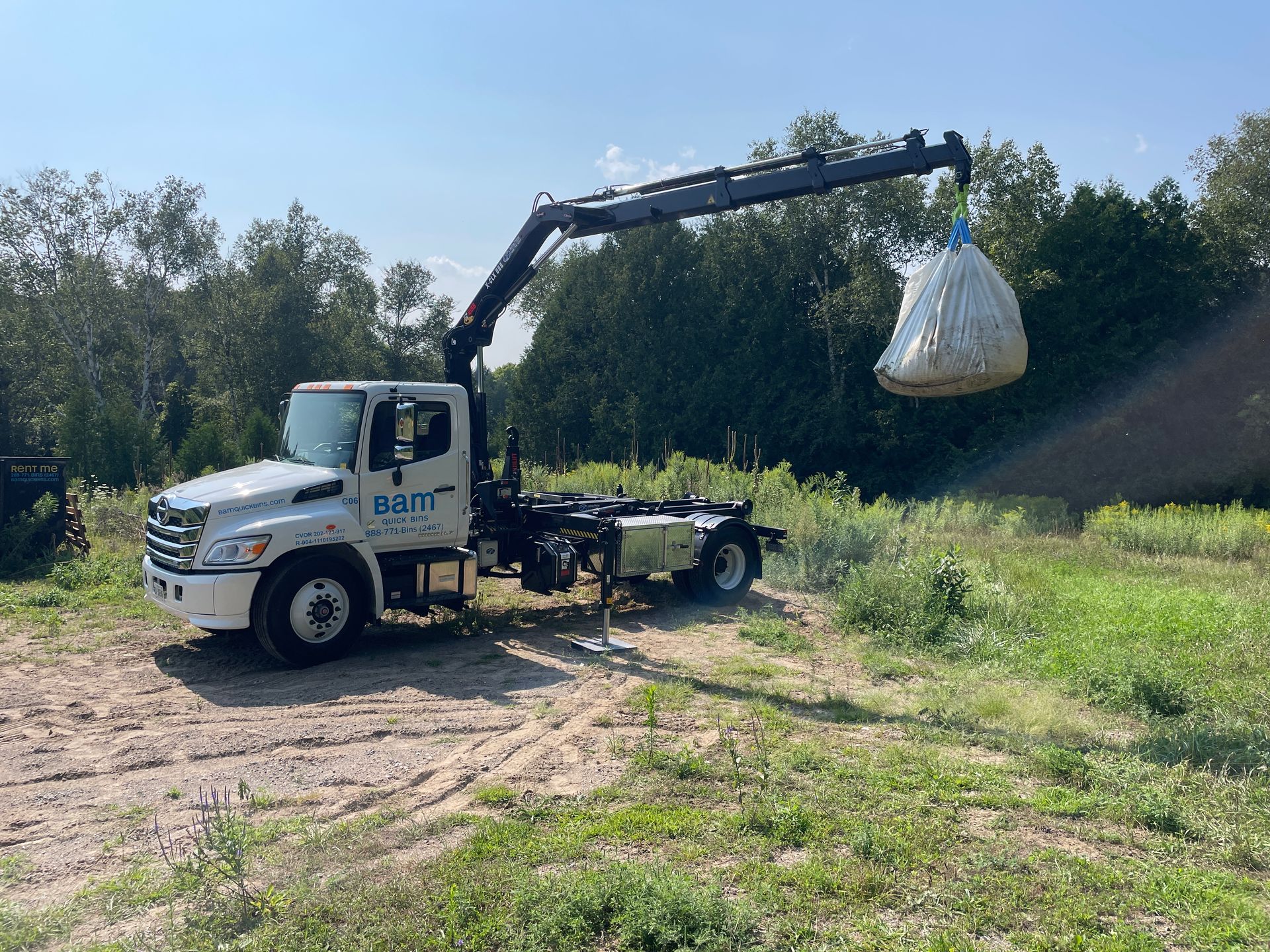 White truck with crane lifting a large bag in a field. Blue sky and trees in the background.