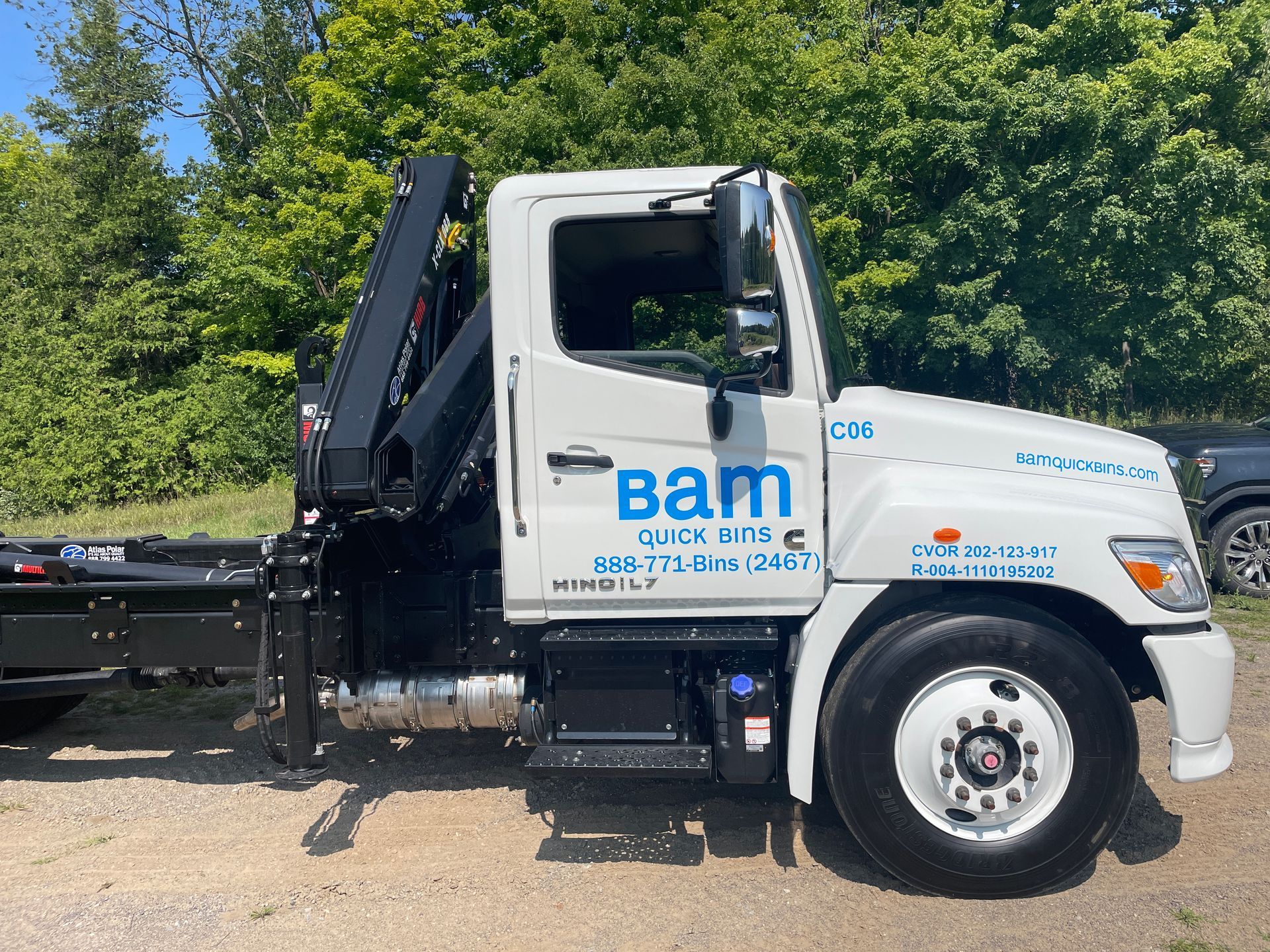 White BAM roll-off truck parked on gravel, with black hoist. Blue logo and contact info on the side.