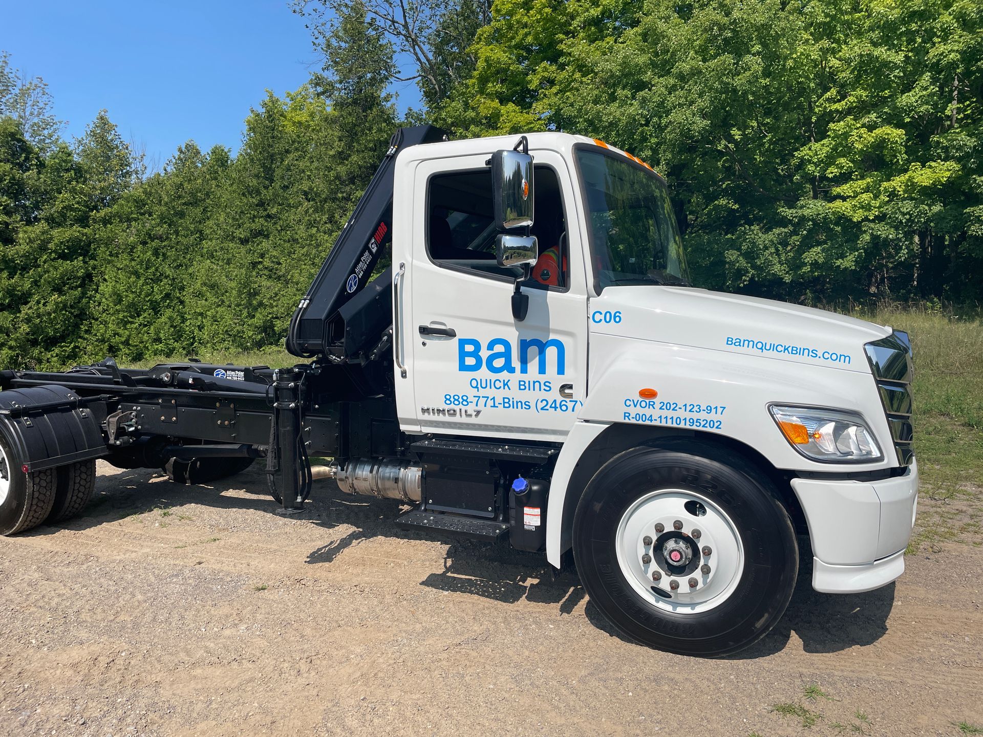 White BAM Waste truck with a black lifting mechanism on a gravel road, trees in the background.