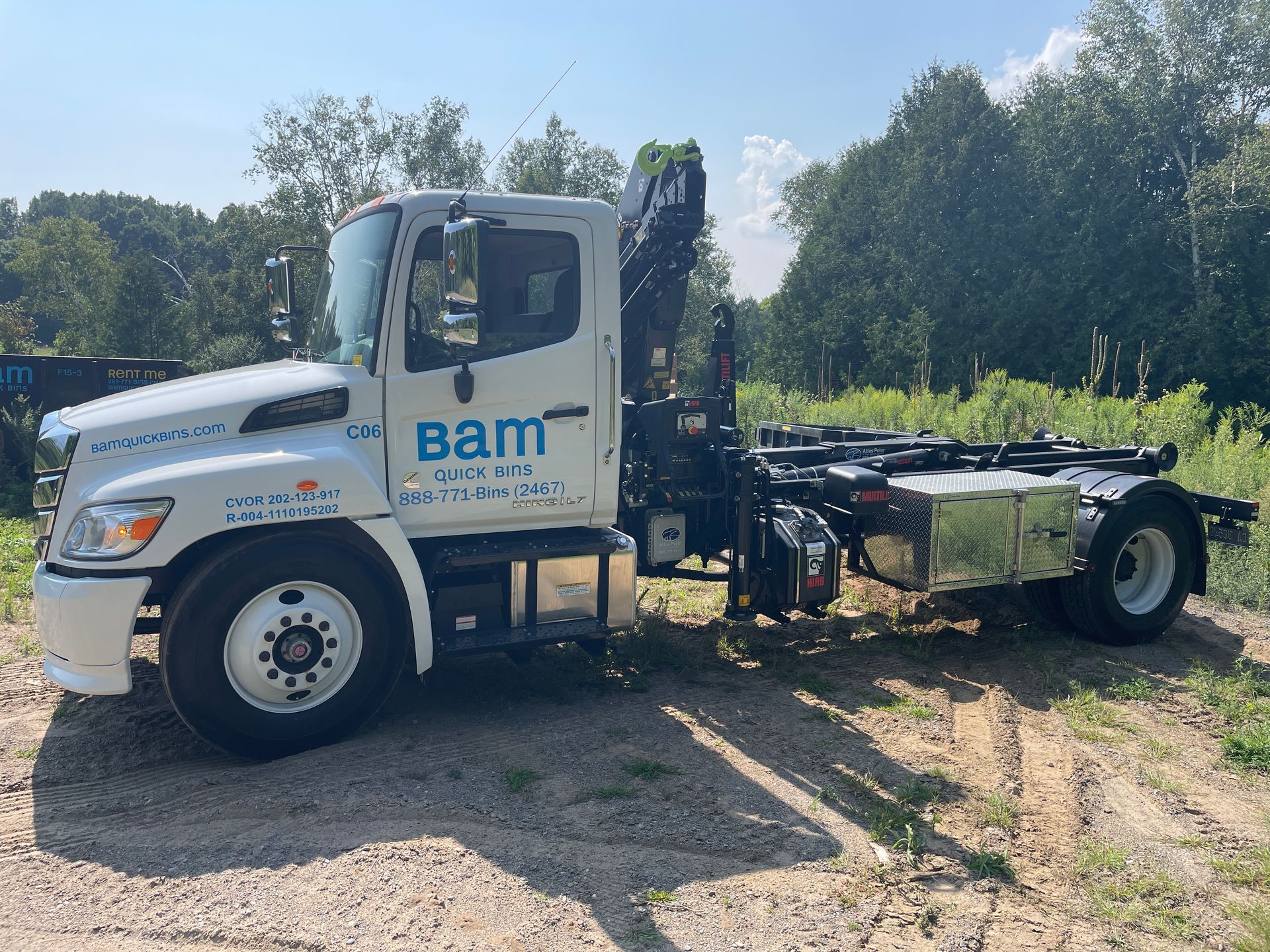 White Bam truck with roll-off container system parked on dirt road, trees in the background.
