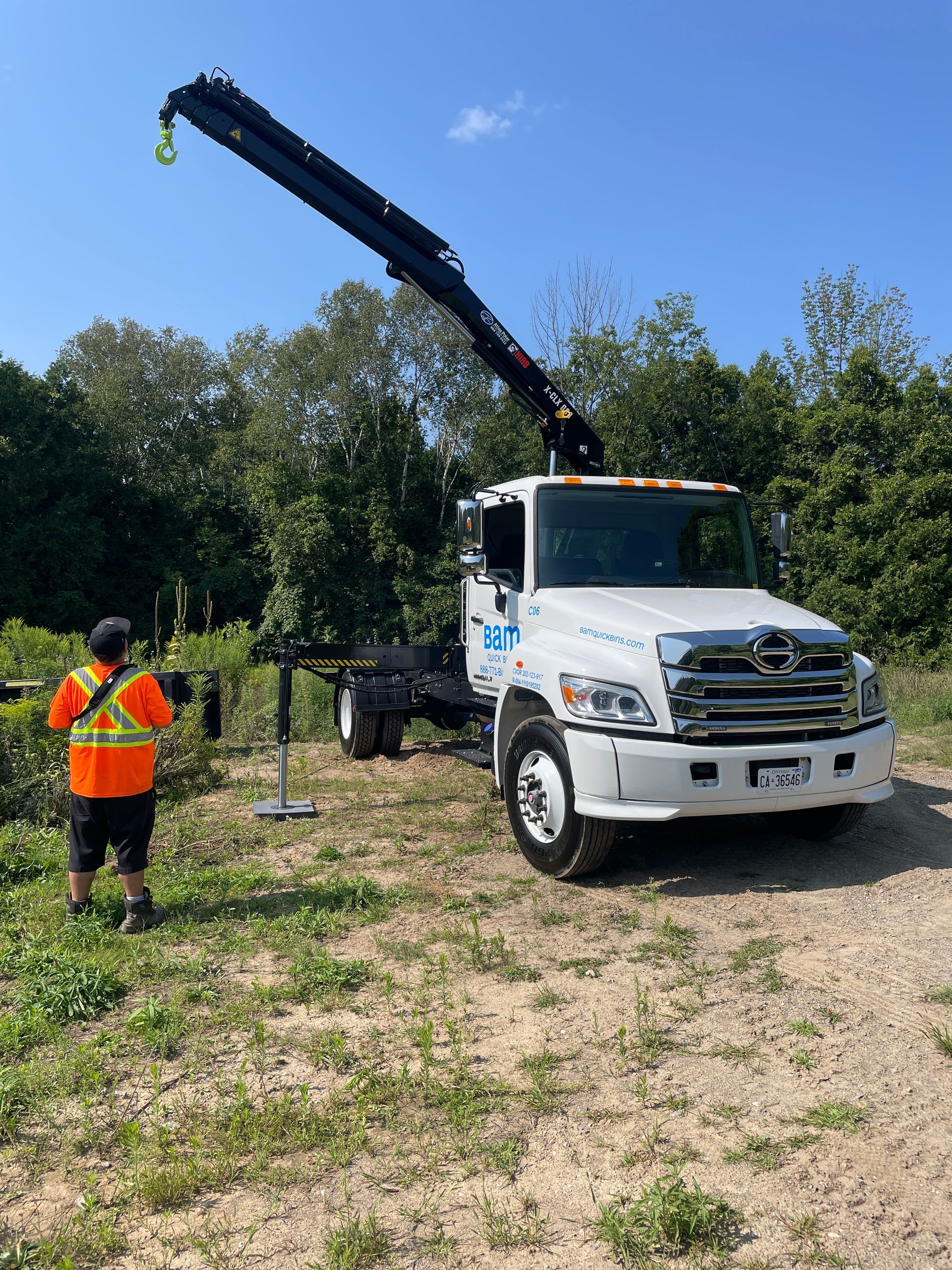 A crane truck with its arm extended, an employee in safety gear stands near, outdoors, blue sky.