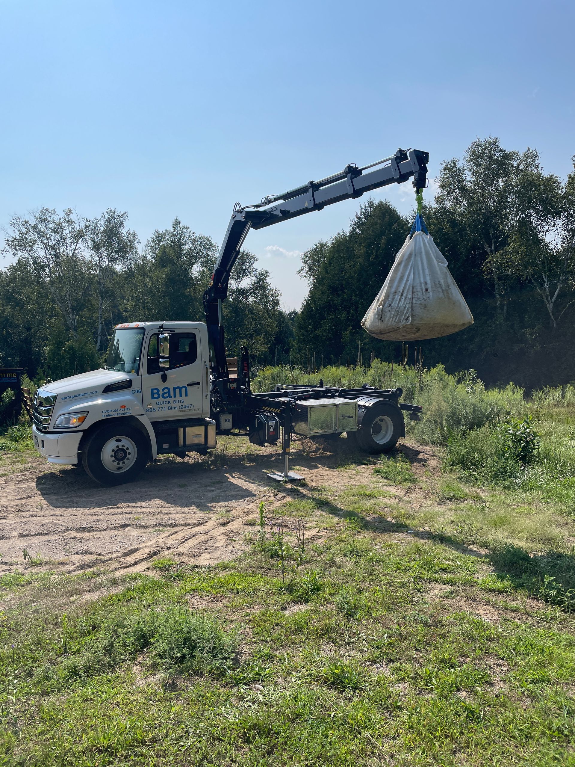 White truck with crane lifting a large bag, outdoors on a sunny day.