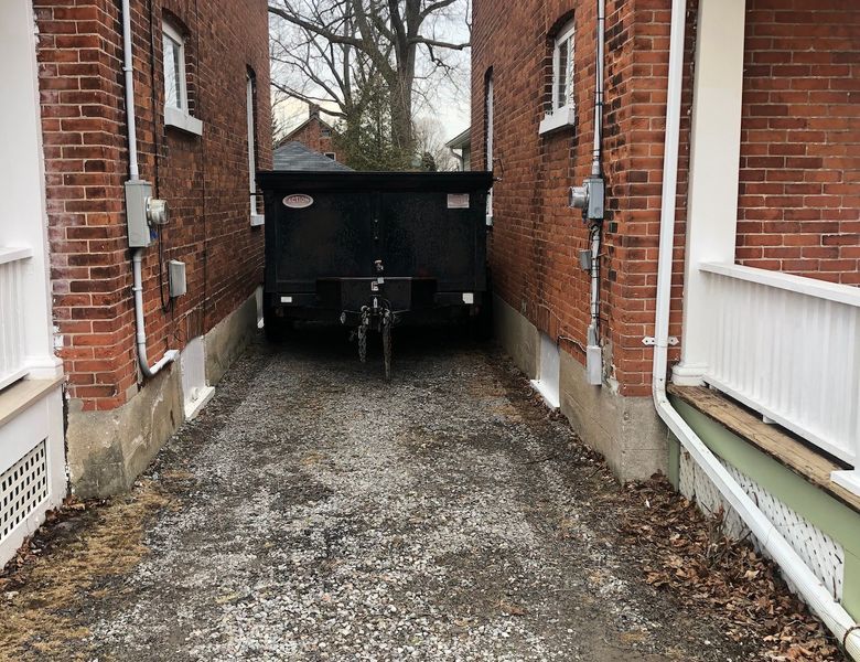 A black dumpster blocks a gravel driveway between two brick buildings.  Gray sky and bare trees are in the background.