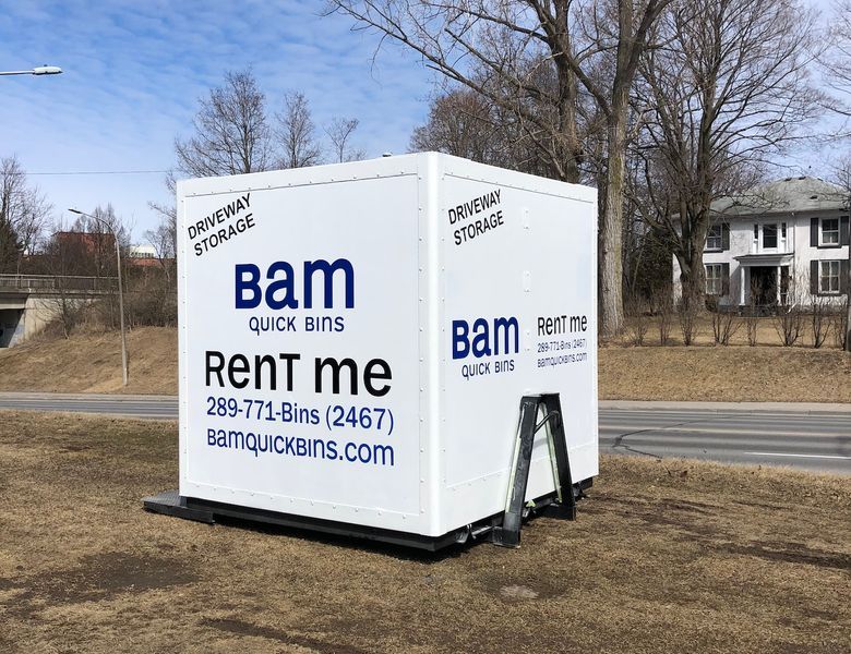 White BAM Quick Bins container for rent, parked on grass near a road. Includes company name, phone number, and website.