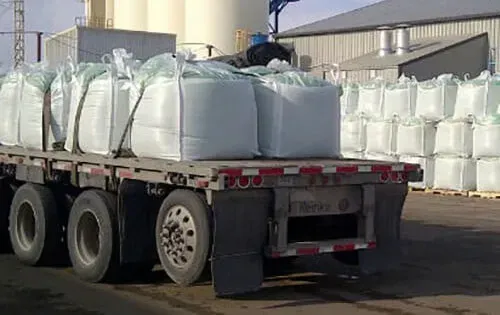 A flatbed truck loaded with large white bags, possibly fertilizer or industrial materials, parked outside a factory.