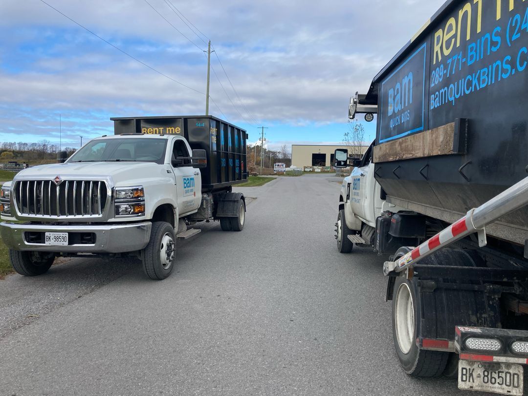 Two white trucks with black dumpsters parked on a gray asphalt road.  The sky is cloudy.