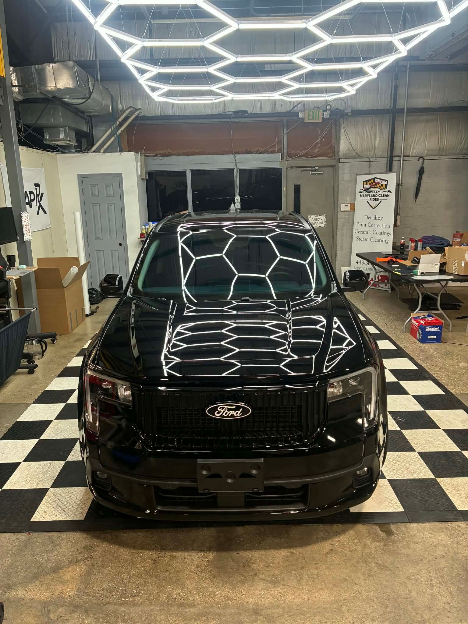 Black SUV parked in a garage with honeycomb lights, reflected on its glossy hood and a checkered floor.