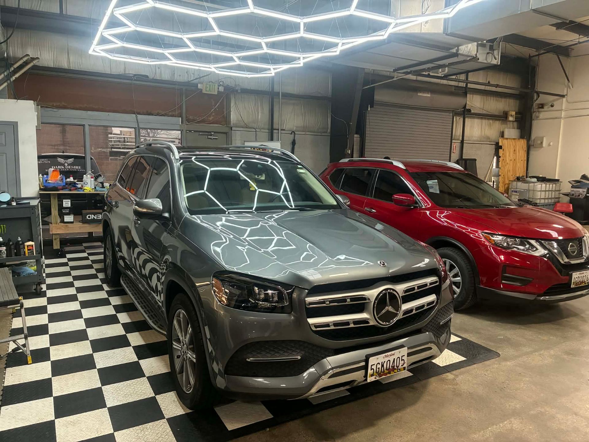 Gray Mercedes SUV and red Nissan SUV parked in a garage with checkered floor. Hexagon lights are overhead.
