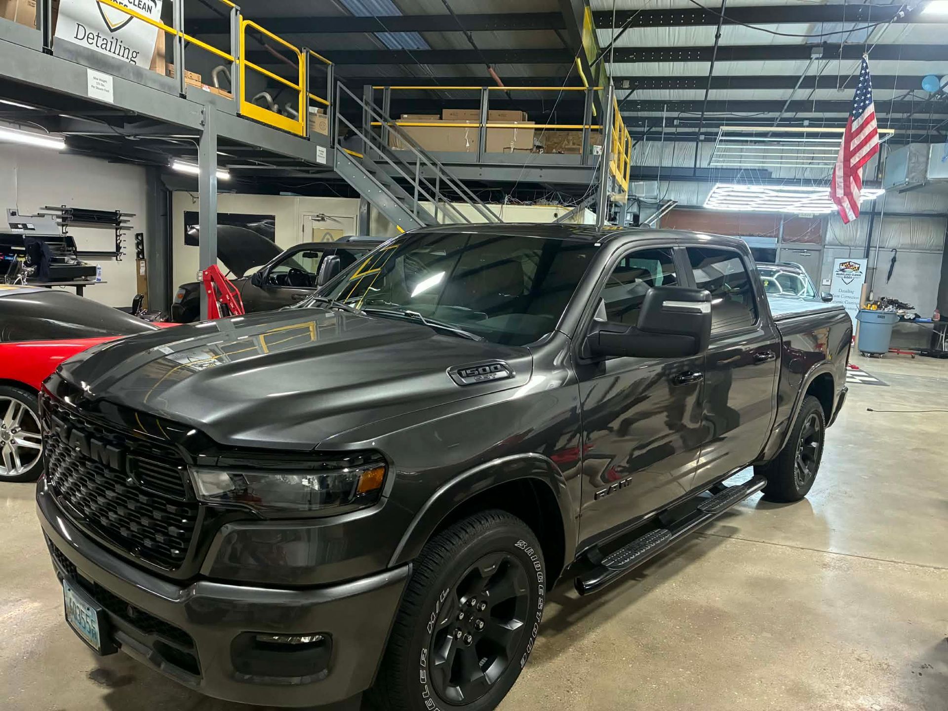 Dark gray Ram pickup truck in a shop with black grille, wheels, and side steps; American flag in the background.