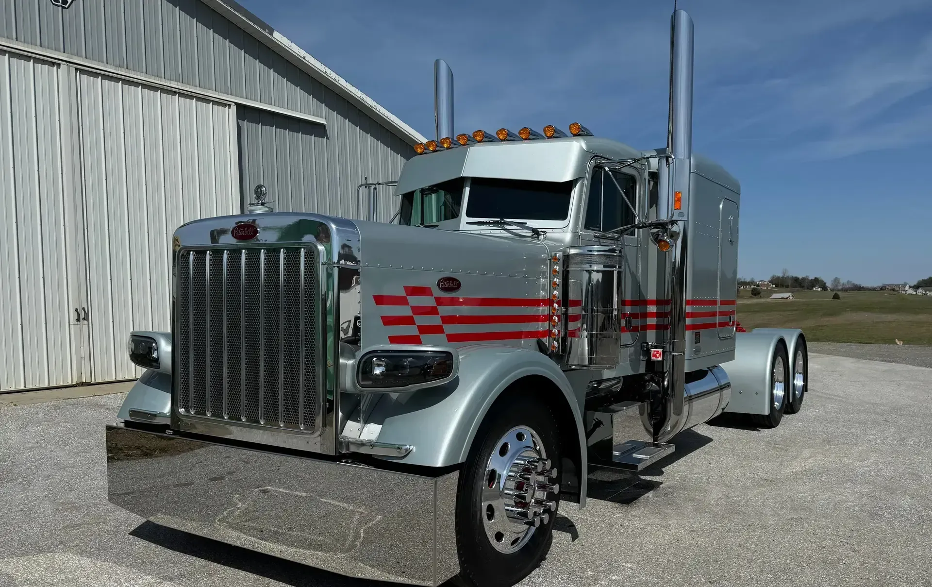 Silver Peterbilt semi-truck with chrome accents and red stripe detailing, parked in front of a barn.