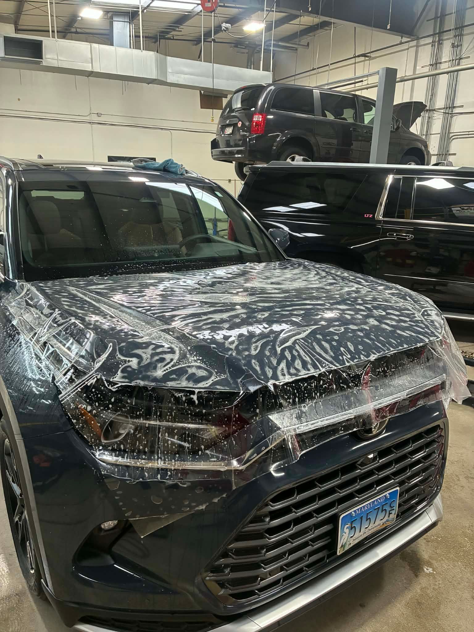 A car's hood covered in protective film, with other vehicles in a garage.