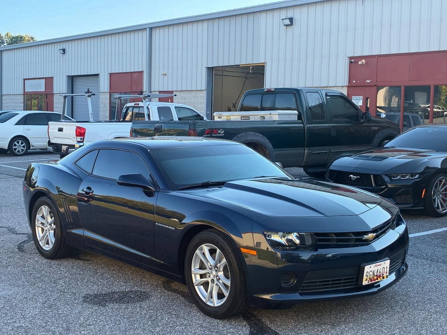 Dark blue Chevrolet Camaro parked in front of a building with other vehicles, gray pavement.