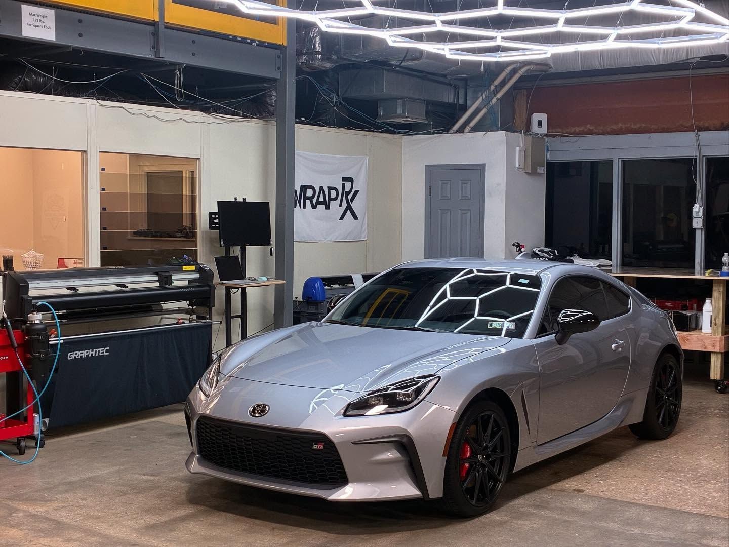 Silver sports car in a shop with hex lights overhead. A banner reads “WRAP FX”.