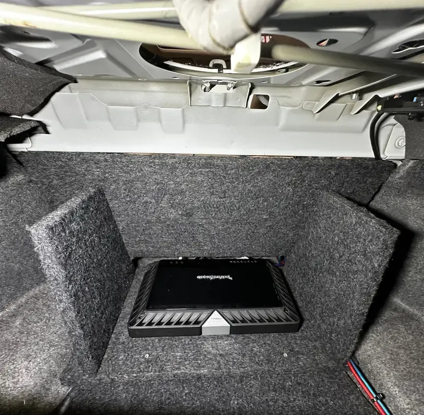 Car audio amplifier installed in a trunk cavity, surrounded by gray carpeting.