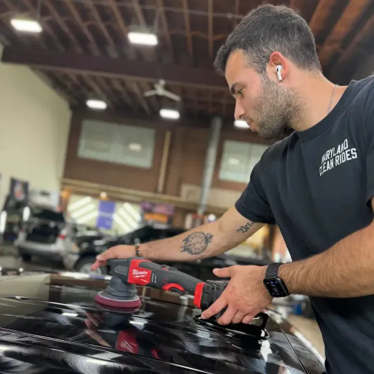A man polishes a black car with a power tool in a garage-like setting.