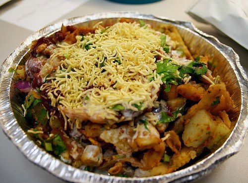 Chopped Indian street food: potatoes, chickpeas, chutney, yogurt, and crispy sev, in a foil container.