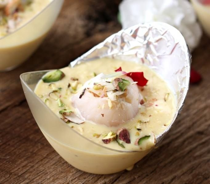 Dessert cup of pale yellow rasmalai, topped with nuts and a pink flower petal, on a wooden surface.