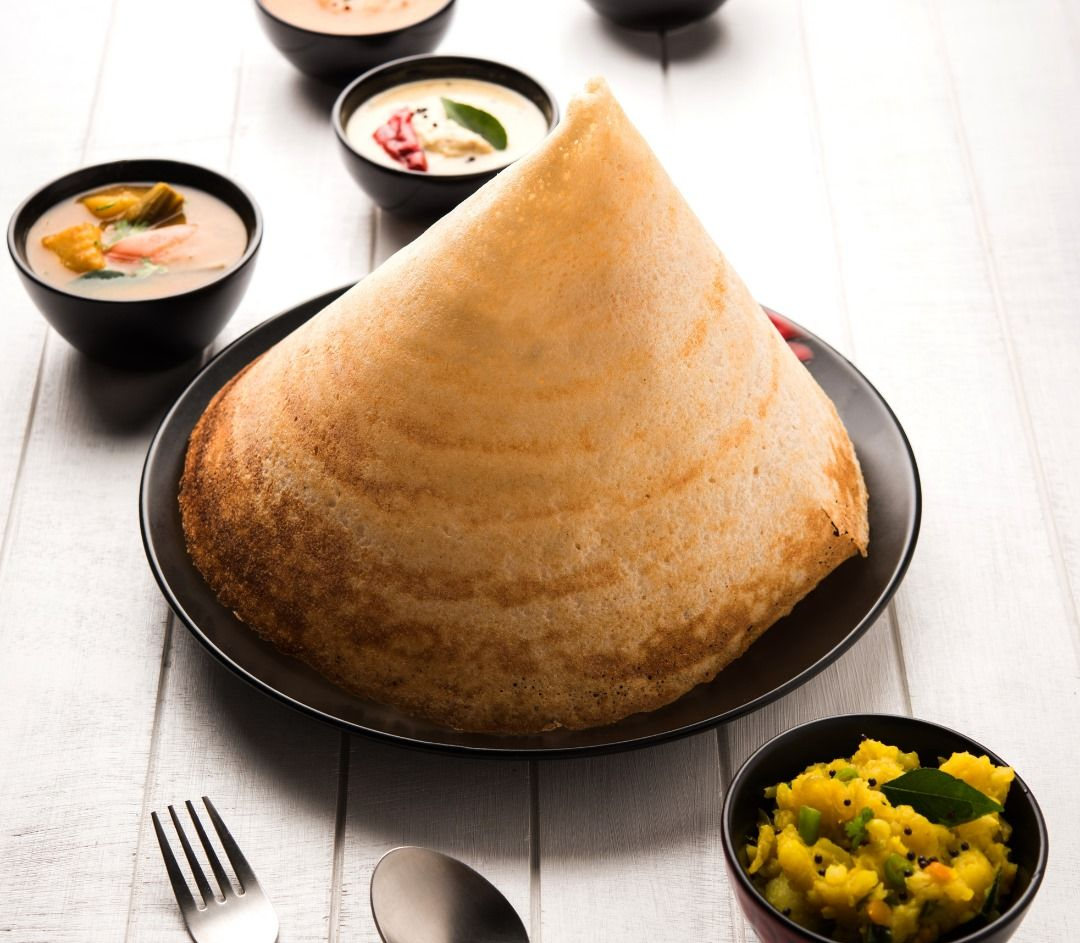 Large conical dosa on a black plate, surrounded by bowls of chutney and curry on a white wooden surface.