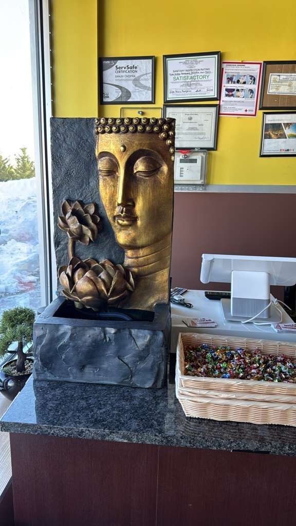 Buddha fountain with gold face, lotus flowers, and a dark stone base, displayed on a counter with awards in the background.