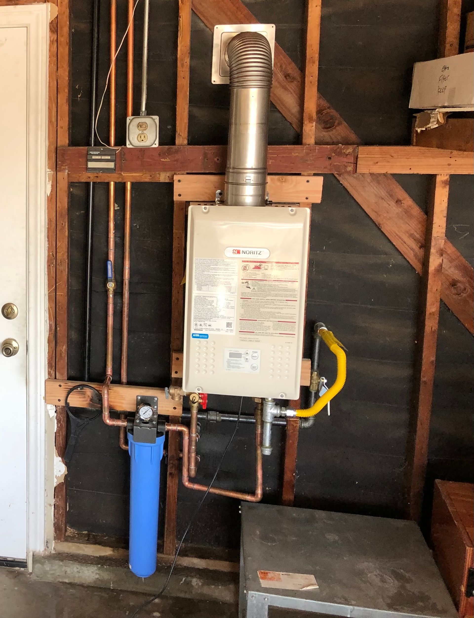 A tankless water heater installed on a garage wall with plumbing, exhaust, and water filter.