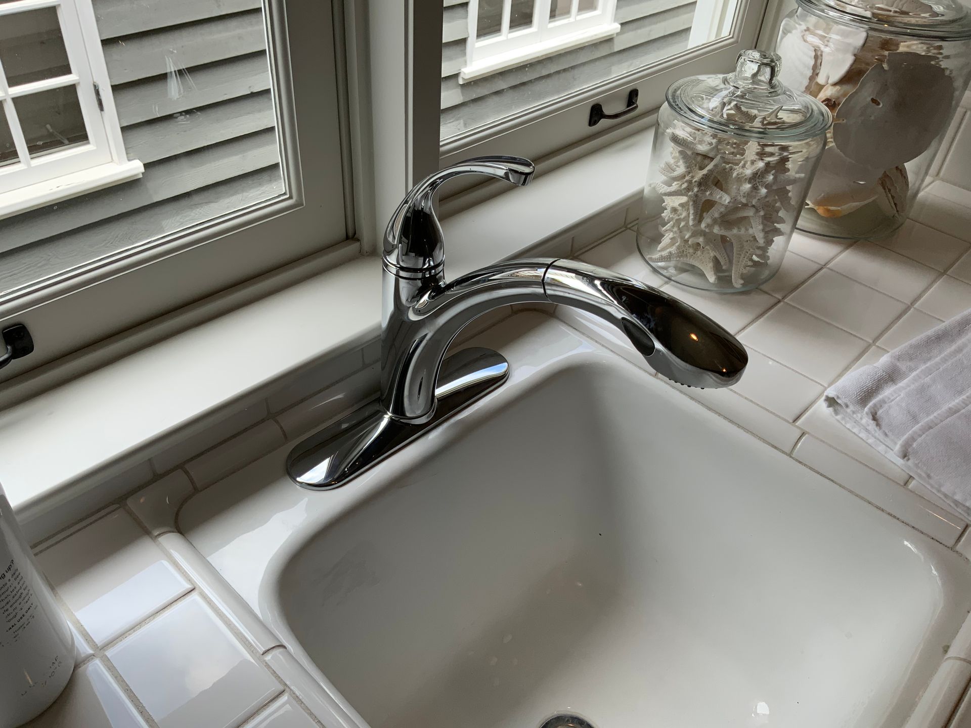 Chrome faucet over a white kitchen sink, near a window and decorative jars.