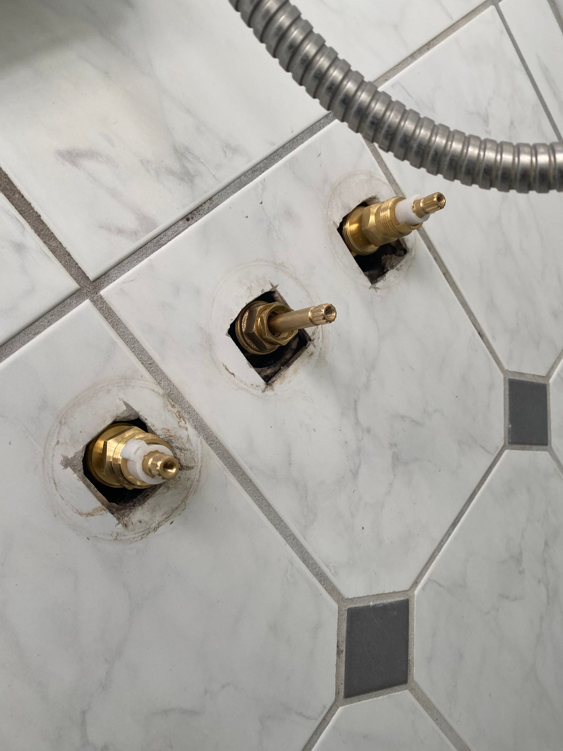 Three brass shower valve stems protrude from white tile. A silver shower hose hangs above.