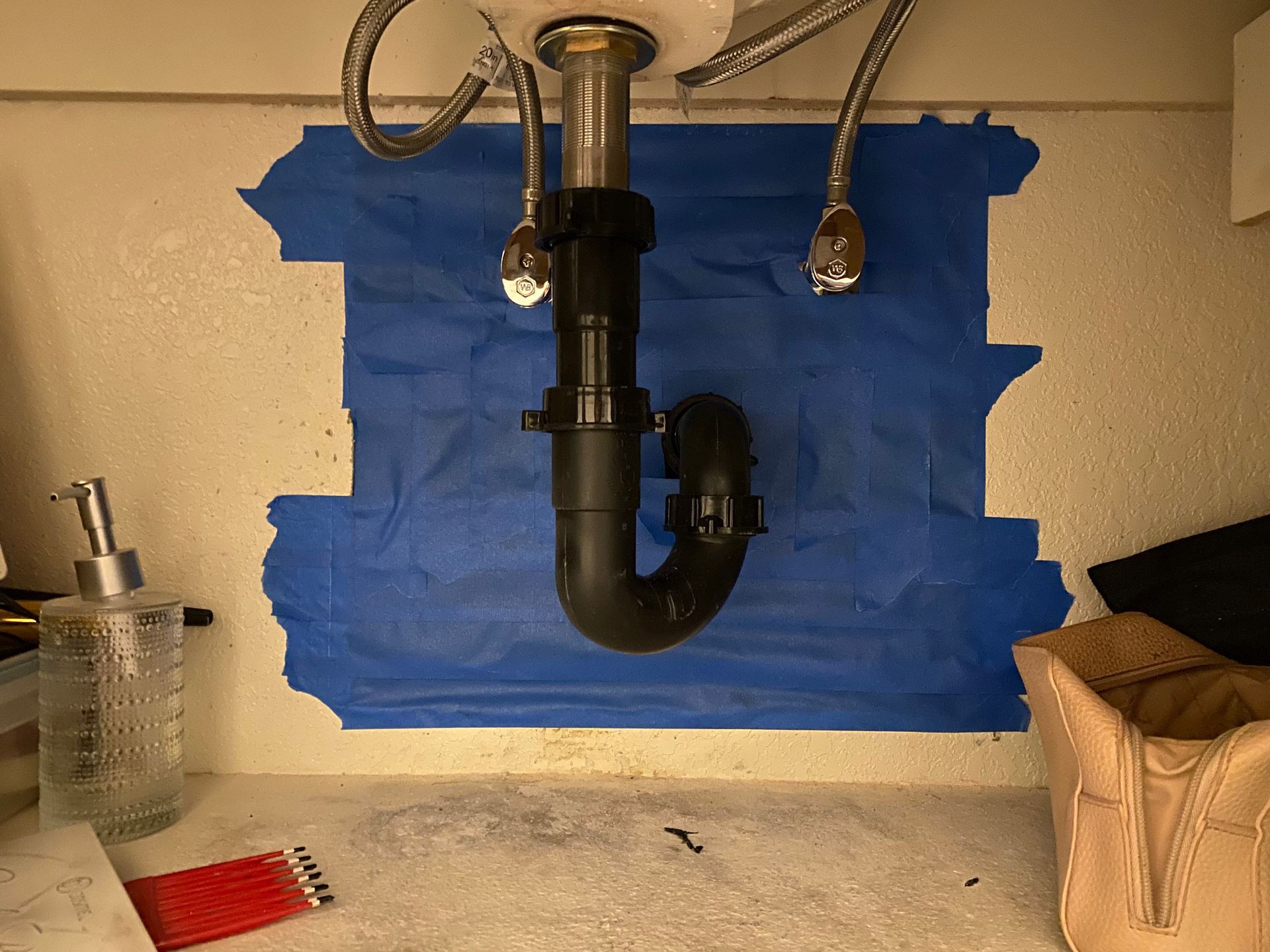 Under-sink plumbing: P-trap, supply lines, and drain, taped area on the cabinet.