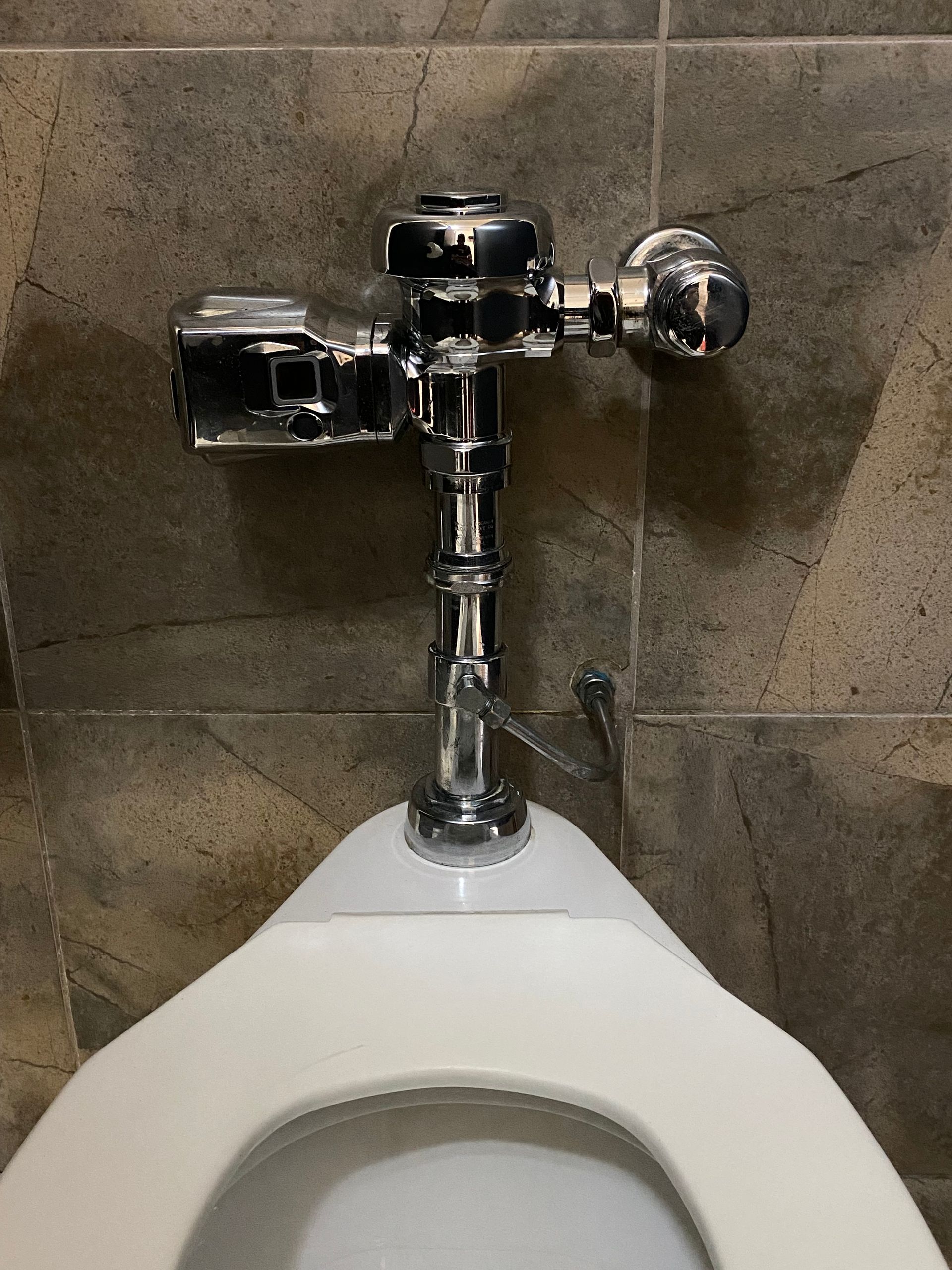 A chrome urinal flush valve atop a white porcelain toilet bowl, against a tiled wall.