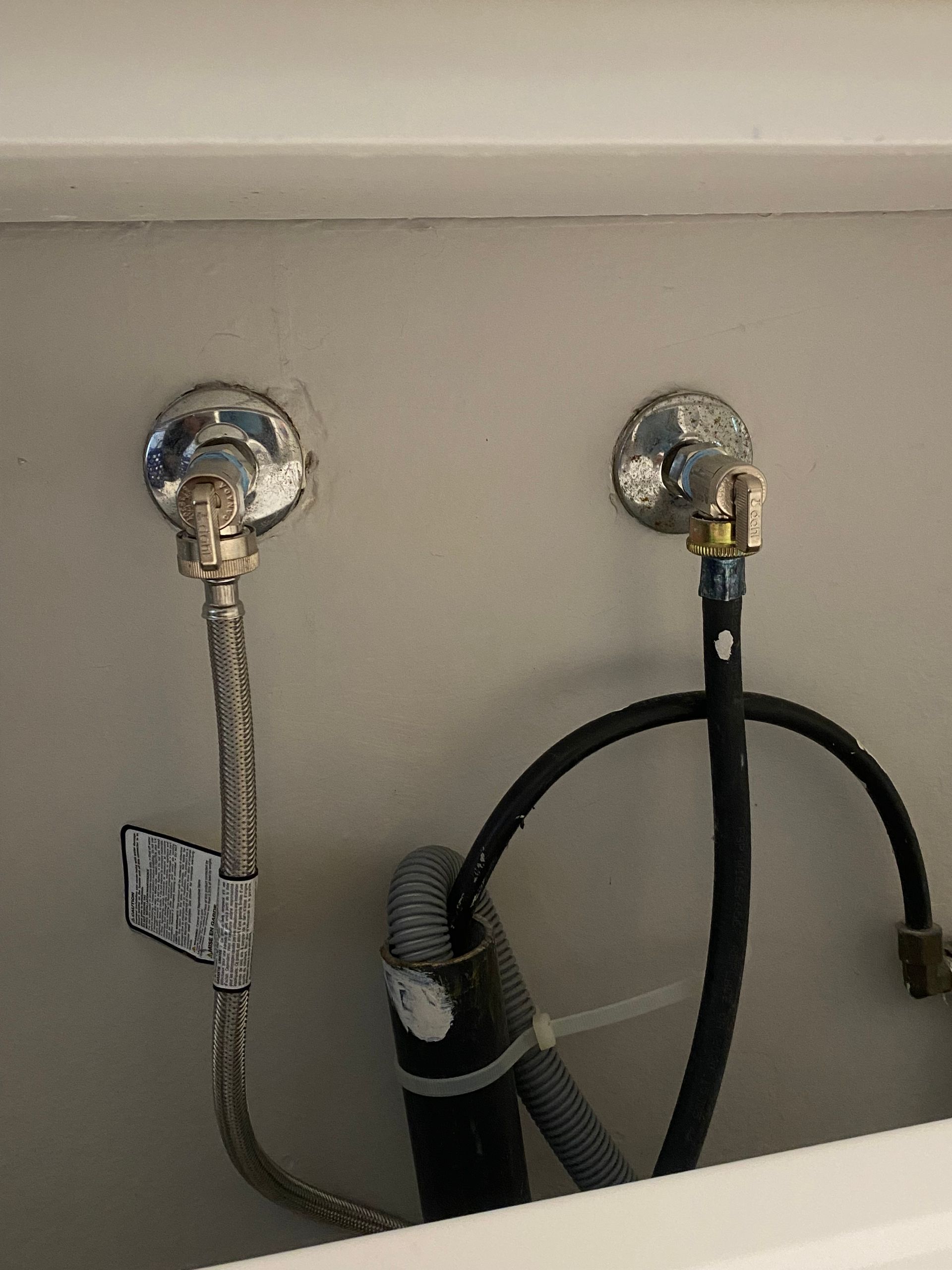 Plumbing under a sink: Chrome shut-off valves, flexible supply lines connected to water pipes on a gray wall.