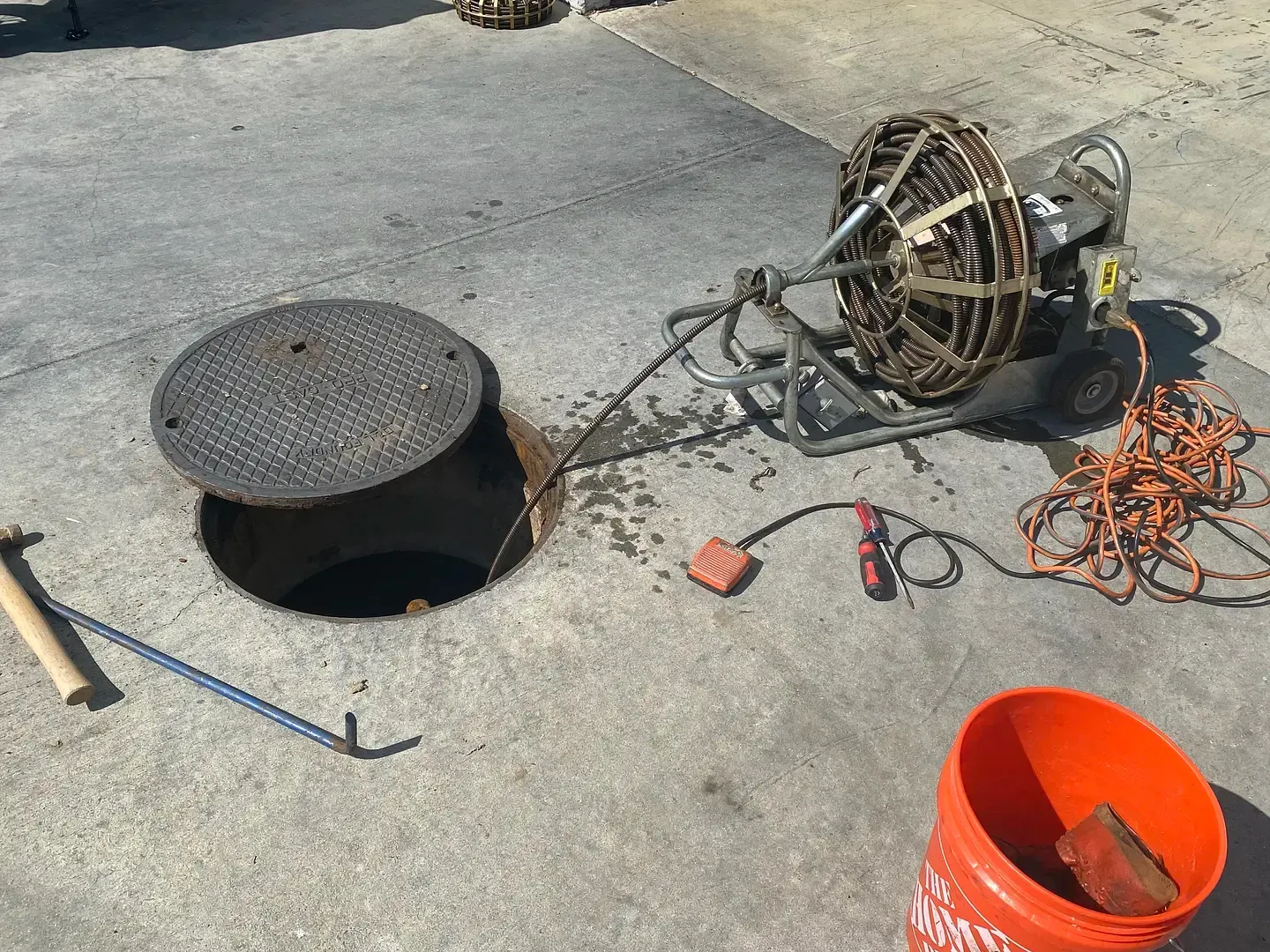 Open manhole with cable machine for drain cleaning; tools and bucket nearby.