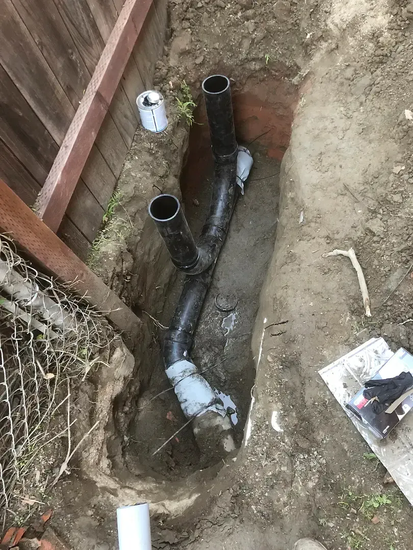 Pipes in an excavated trench, likely for plumbing, near a fence in a yard.