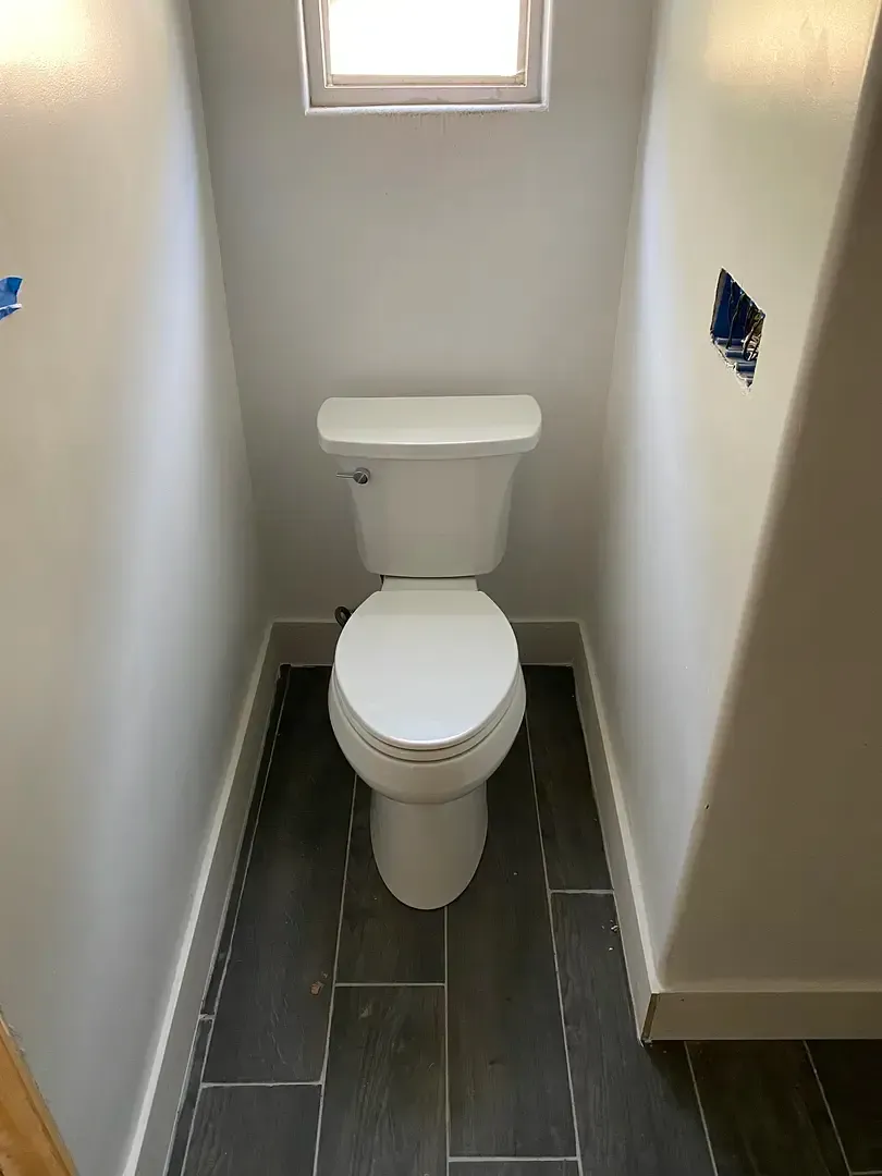 White toilet in a small, narrow bathroom with gray tile flooring and a small window above.